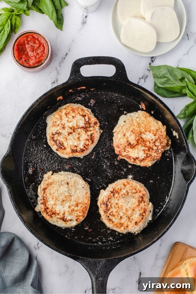 Ground chicken burgers cooking in a cast iron skillet, topped with melting mozzarella