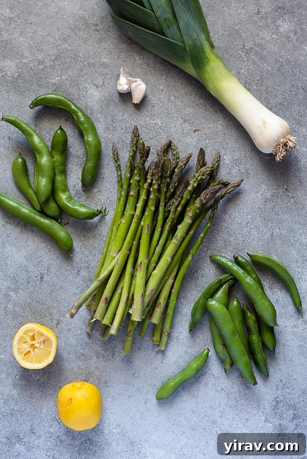 Vibrant Spring Risotto 4 Ingredients laid out on a white surface: fresh asparagus, shelled fava beans, sliced leeks, garlic cloves, lemons, a block of parmesan cheese, and arborio rice.
