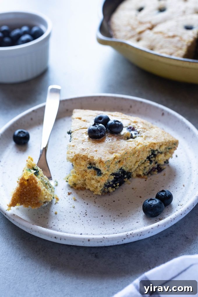 A perfectly sliced piece of blueberry cornbread on a white plate, with a fork resting beside it, highlighting its fluffy texture and visible blueberries.