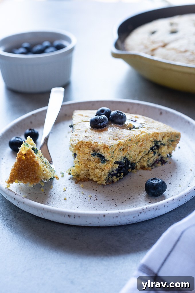A close-up image of a wedge of blueberry cornbread on a white plate, with a bite taken out, showing the soft, fluffy interior and visible blueberries.