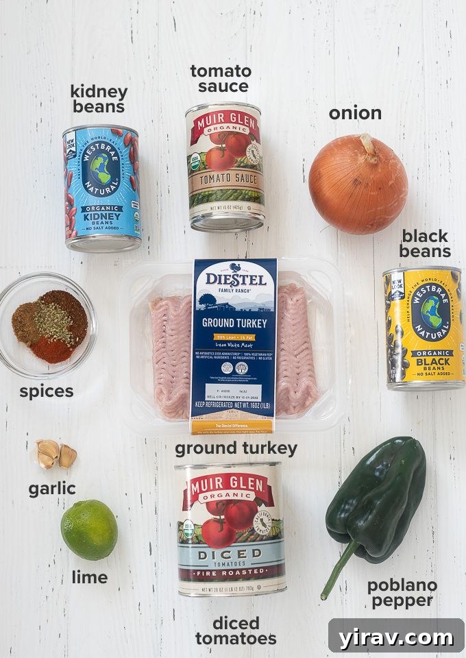 All the essential ingredients for slow cooker turkey chili laid out on a kitchen counter: ground turkey, canned tomatoes, beans, onions, garlic, poblano pepper, and various spices.