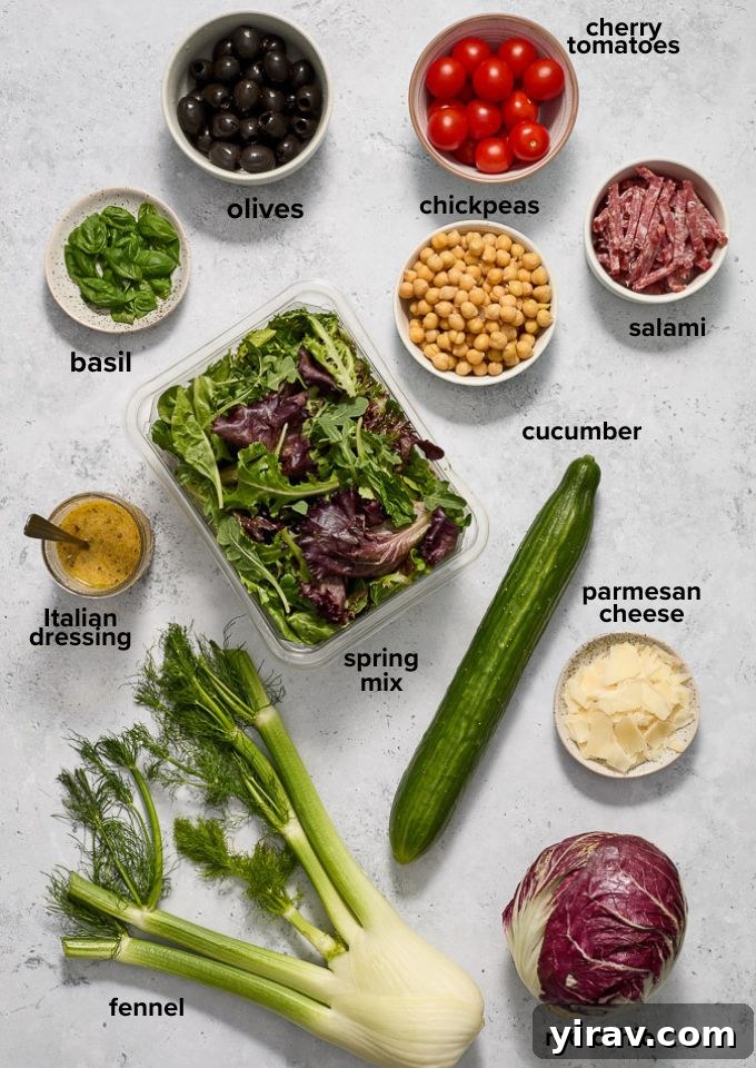 Various fresh ingredients for Italian chopped salad, including greens, tomatoes, cucumber, salami, olives, and chickpeas.