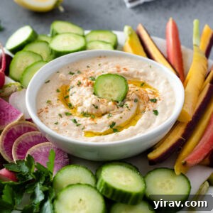 Creamy White Bean Hummus 8 A bowl of creamy white bean hummus garnished with a drizzle of olive oil and fresh herbs, surrounded by colorful vegetable sticks for dipping.