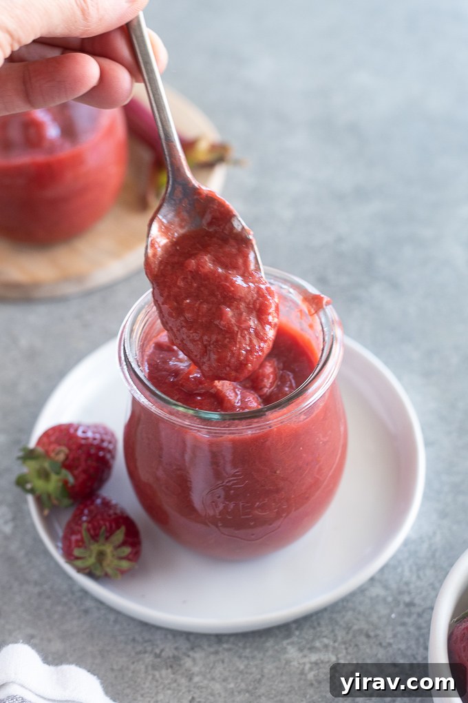 A spoonful of homemade rhubarb and strawberry jam held over a jar, highlighting its glistening texture and a few fruit pieces.