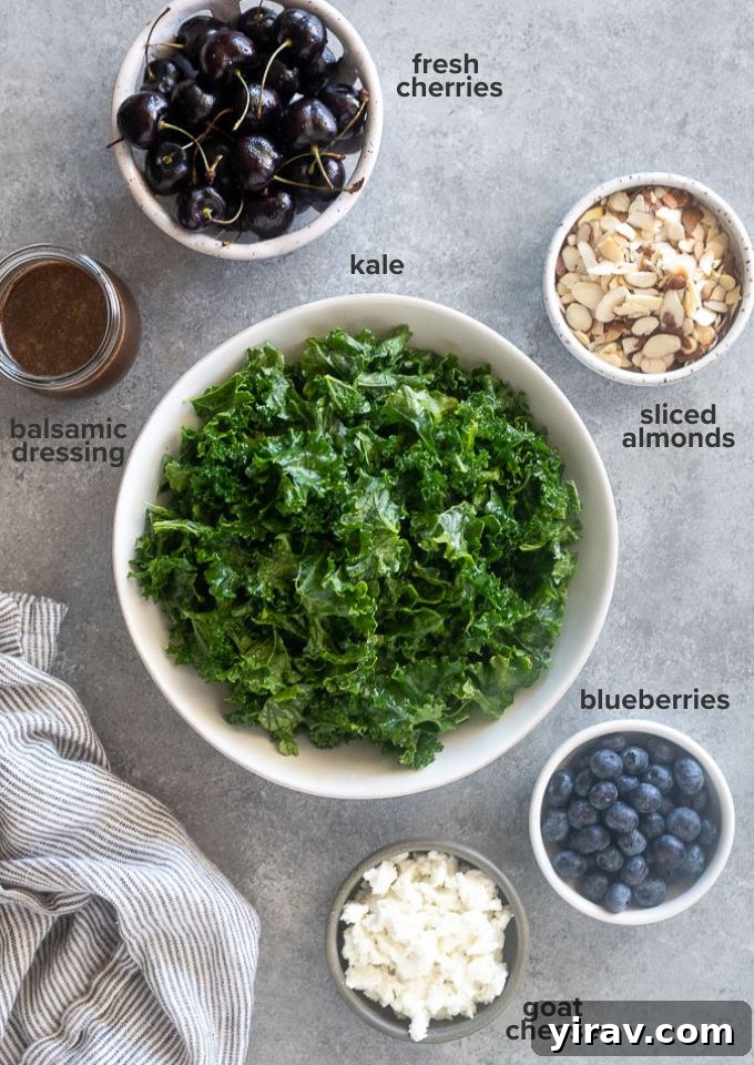 Individual ingredients for Kale Cherry Salad laid out on a white surface, including kale, cherries, blueberries, goat cheese, and almonds