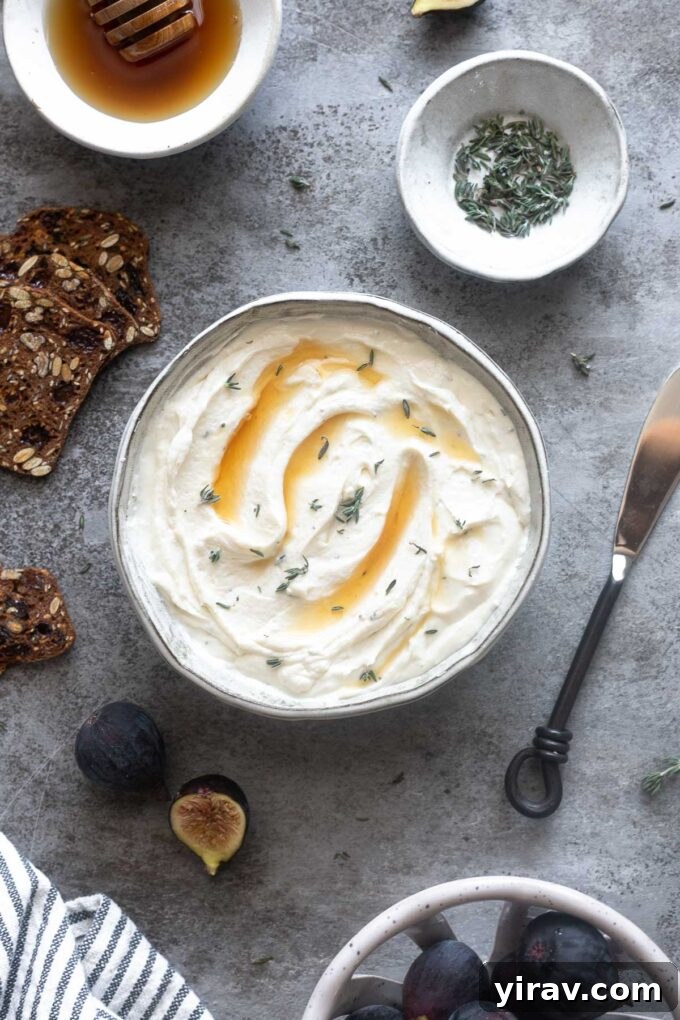A vibrant serving of whipped goat cheese in a white bowl, garnished with honey and fresh thyme, surrounded by an arrangement of fresh figs and crackers.