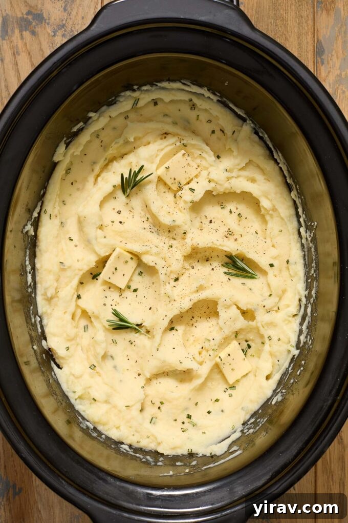 Close-up of buttermilk mashed potatoes in a crock pot insert, garnished with rosemary and melting butter.