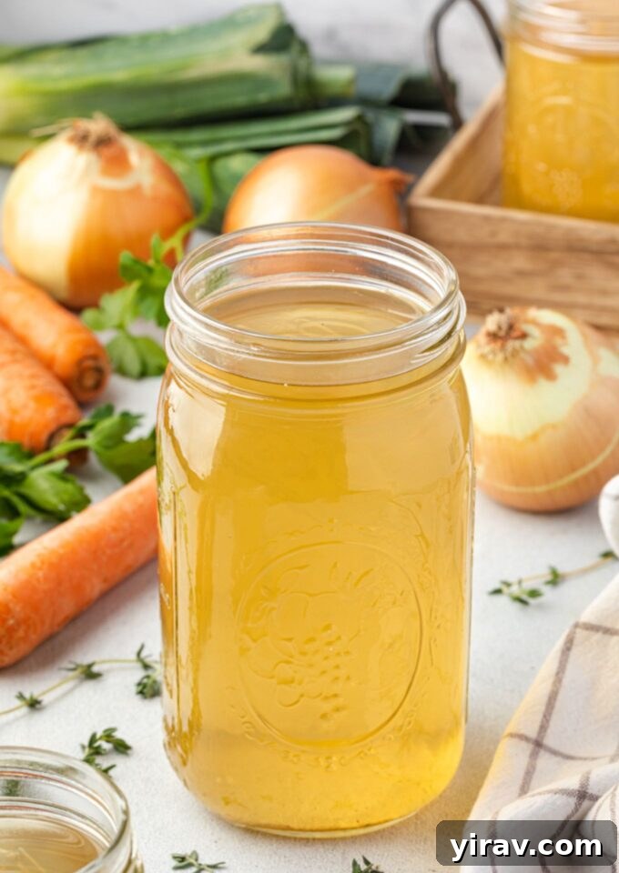 A glass jar filled with golden homemade vegetable broth, surrounded by fresh vegetables like carrots, celery, onions, and herbs, suggesting a healthy and natural cooking process.