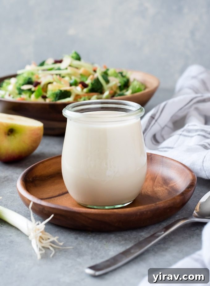 A beautifully presented glass jar brimming with creamy homemade coleslaw dressing, with a refreshing broccoli salad blurred elegantly in the background. The image highlights the dressing's smooth texture and appetizing appearance, suggesting its perfect pairing with fresh salads.