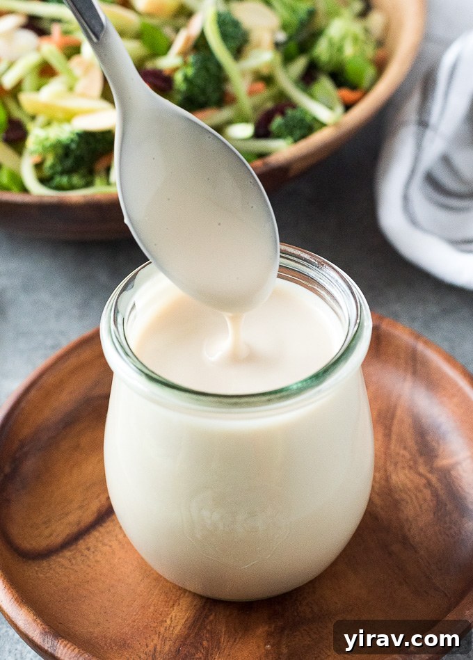 A hand holding a spoon, gracefully drizzling rich, creamy homemade coleslaw dressing into a glass jar. The motion beautifully illustrates the dressing's smooth, pourable consistency, making it look irresistible and ready to use.