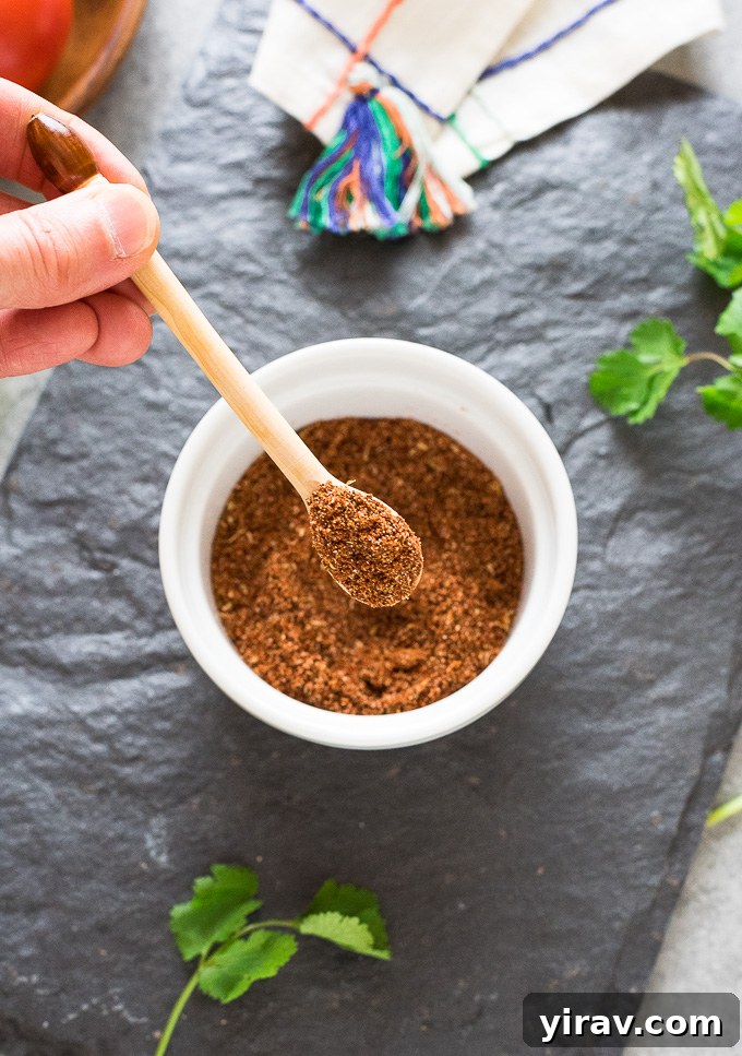 A close-up of a spoonful of rich, red-brown Mexican spice mix, held over a glass bowl, showcasing its finely ground texture and vibrant color.