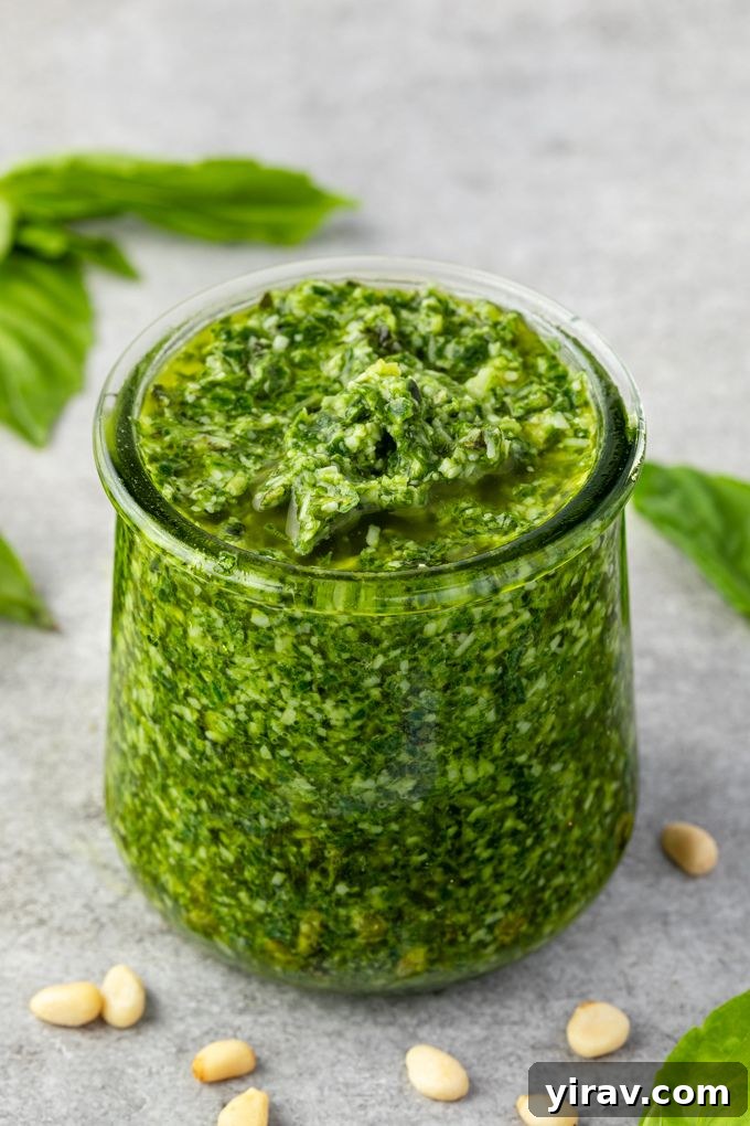 Fresh basil pesto in a clear jar surrounded by pine nuts and fresh basil leaves on a white surface