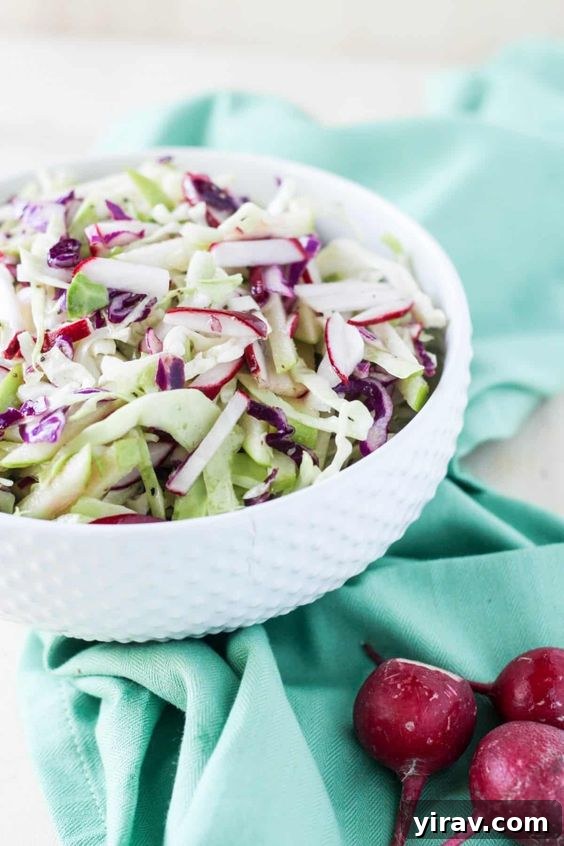 A white bowl of cabbage, radish, and apple coleslaw on a blue towel, vibrant and fresh.