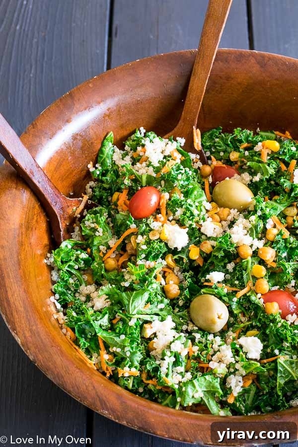 A wooden bowl full of vibrant Roasted Corn and Chickpea Kale Salad, accompanied by two wooden serving spoons.