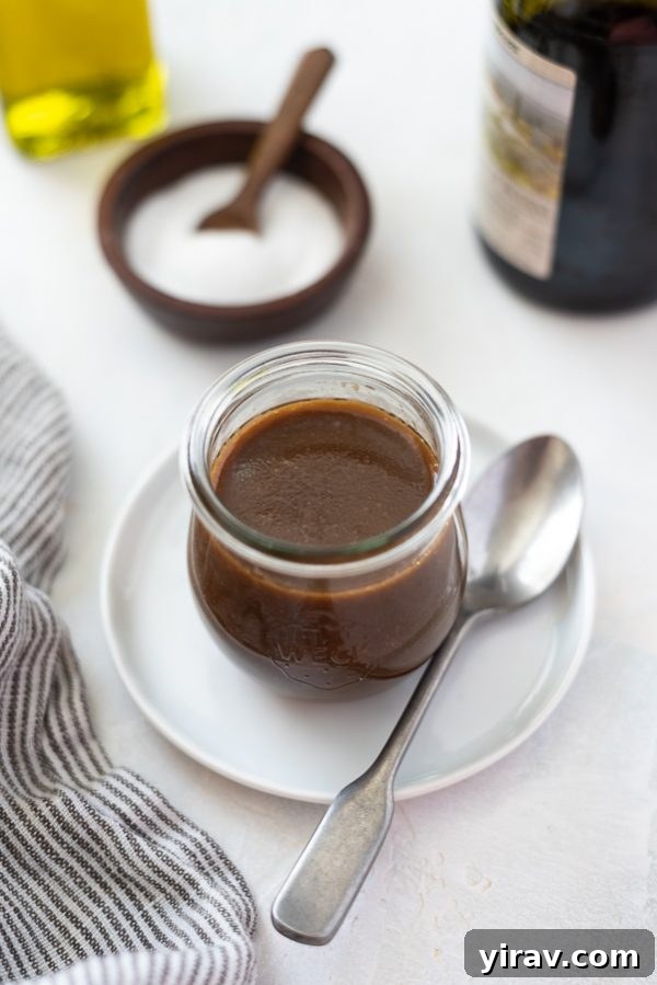 Overhead shot of freshly made balsamic vinaigrette dressing in a clear glass jar, with a serving spoon beside it, on a light background.