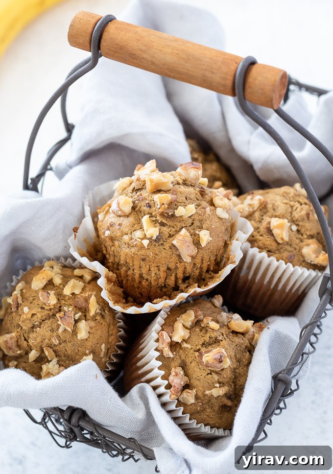 A healthy banana oatmeal muffin with its paper liner partially removed, showing its soft, moist interior, surrounded by other muffins in a woven basket.