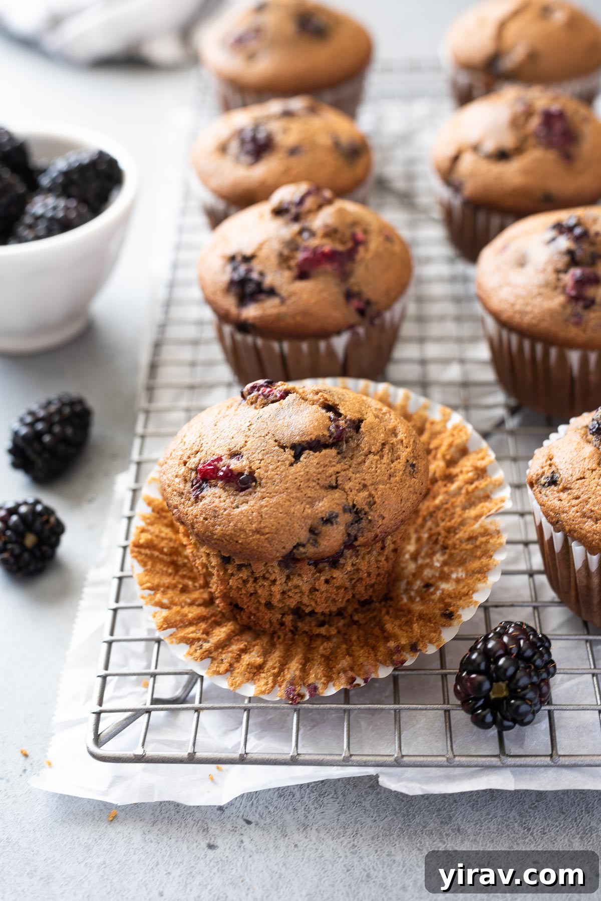 A single perfectly baked whole grain blackberry muffin on a wire rack, with its paper liner gently peeled back to reveal its tender interior and juicy fruit.