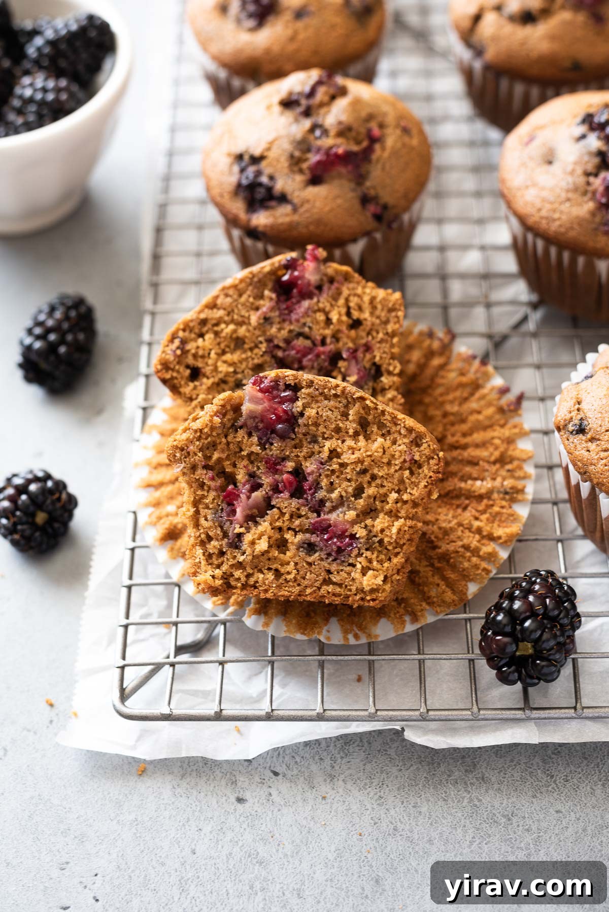 A healthy whole grain blackberry muffin sliced in half, revealing the fluffy interior and vibrant juicy blackberries, sitting on its paper liner on a wire rack.