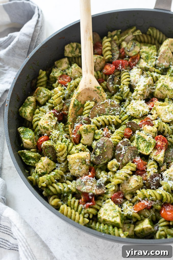 Wooden spoon scooping chicken pesto pasta in a skillet, showcasing the saucy texture.