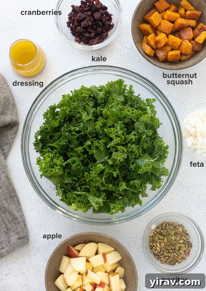 Various fresh ingredients for a harvest salad laid out on a wooden board, including a bunch of kale, chopped butternut squash, a red apple, crumbled feta cheese, dried cranberries, and pepitas, all ready for preparation.