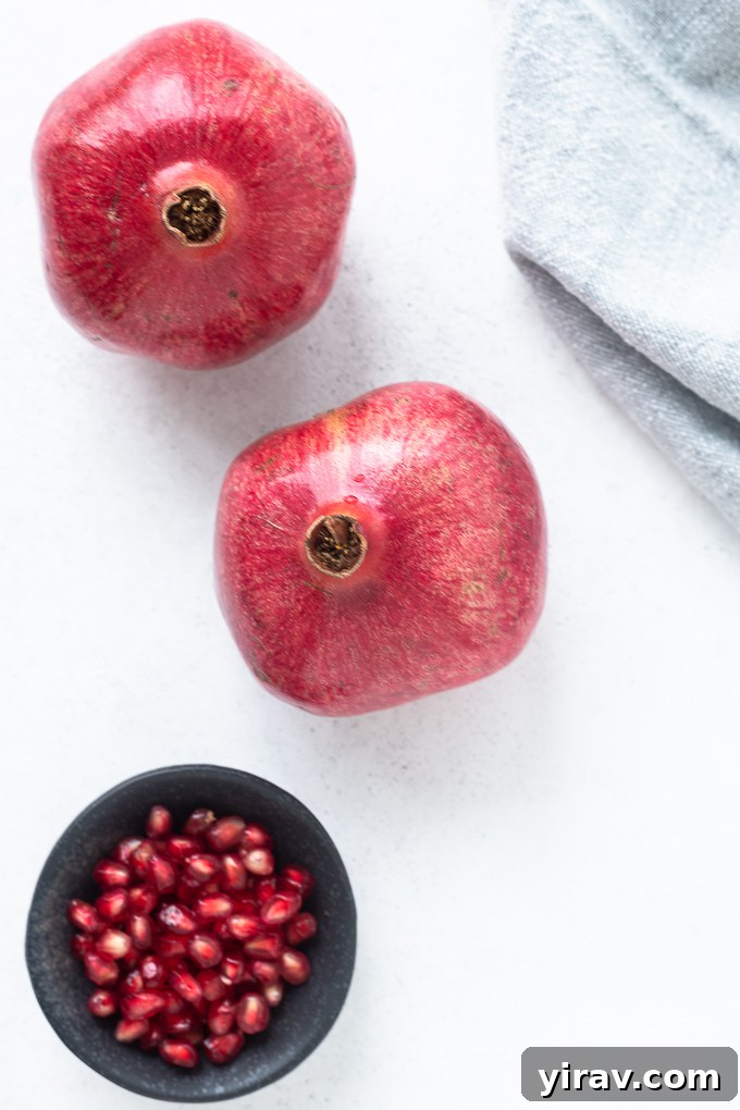 Two whole pomegranates placed next to a clear bowl filled with glistening red arils.