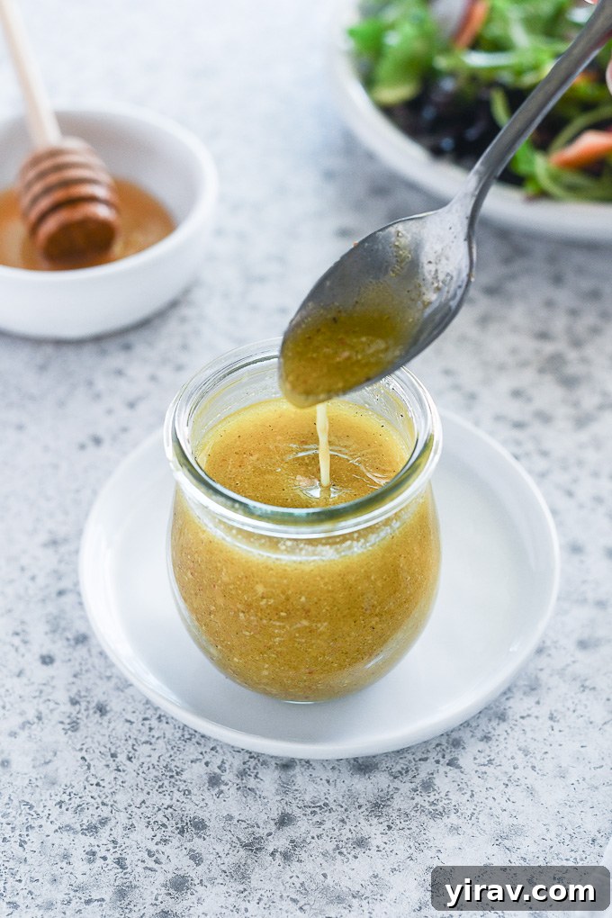 A spoon expertly drizzling thick, golden honey mustard dressing into a glass jar.
