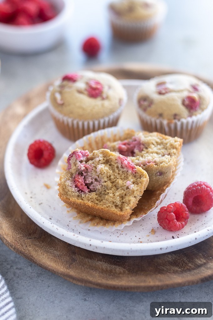 A close-up of a raspberry muffin cut in half, showcasing the soft interior and evenly distributed raspberries on a white plate.