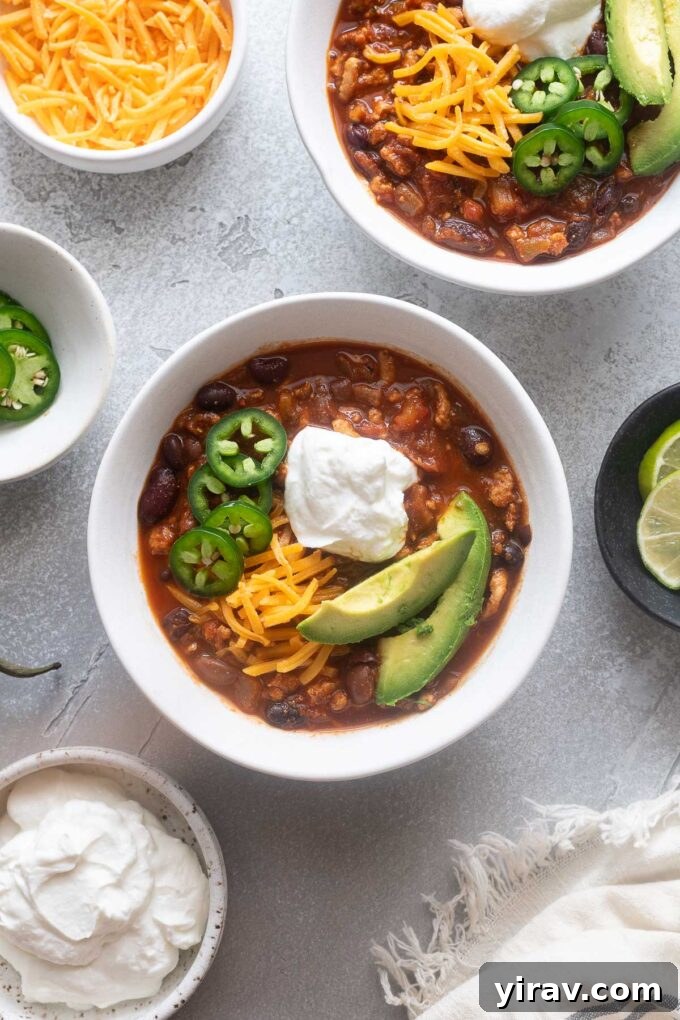 Ground chicken chili in a bowl surrounded by various toppings like cheese, avocado, and lime.
