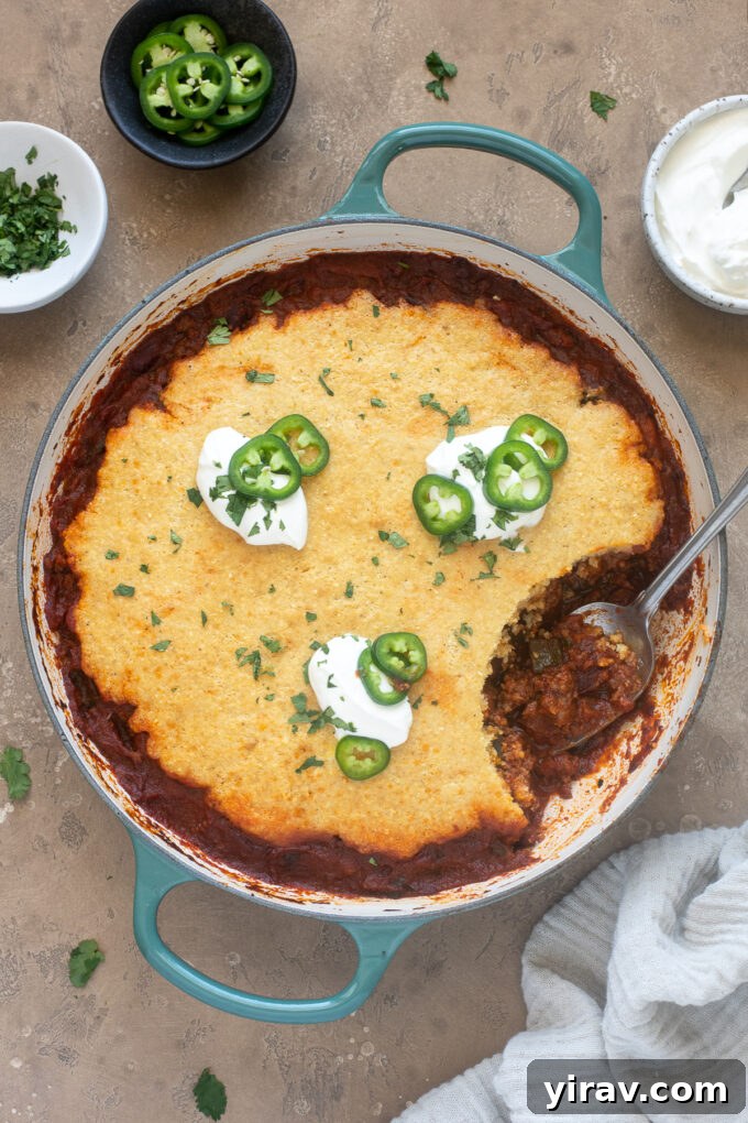 Comforting Chili Cornbread Bake 2 Chili cornbread casserole in a cast iron pan with a spoon scooping a portion, showcasing the rich chili and fluffy cornbread layers.