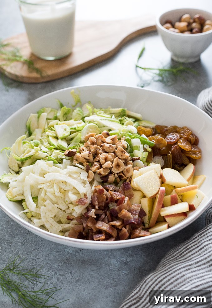 A vibrant flat lay image showcasing all the fresh ingredients for Shaved Brussels Sprout Salad including whole Brussels sprouts, bacon, fennel, apples, raisins, and hazelnuts.