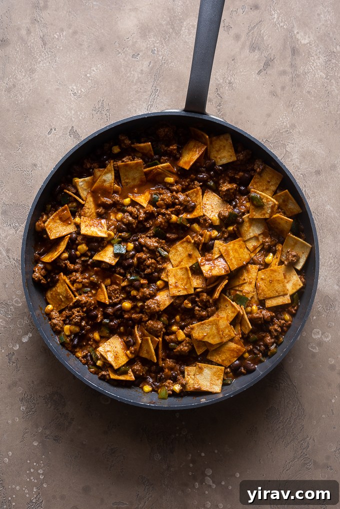 Adding red enchilada sauce, black beans, corn, and cut corn tortillas to the cooked ground beef mixture in the skillet.