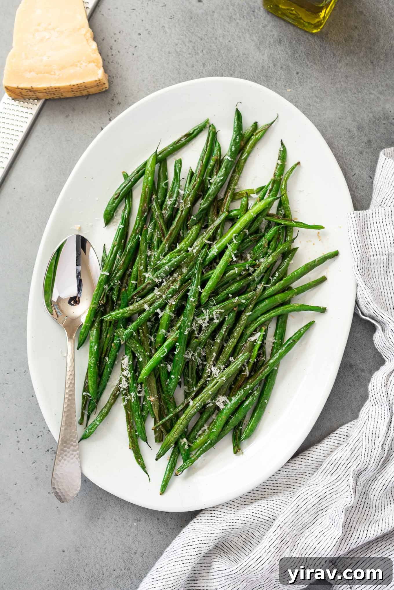 A platter of beautifully roasted green beans, sprinkled with Parmesan cheese, accompanied by a serving spoon and a linen napkin.