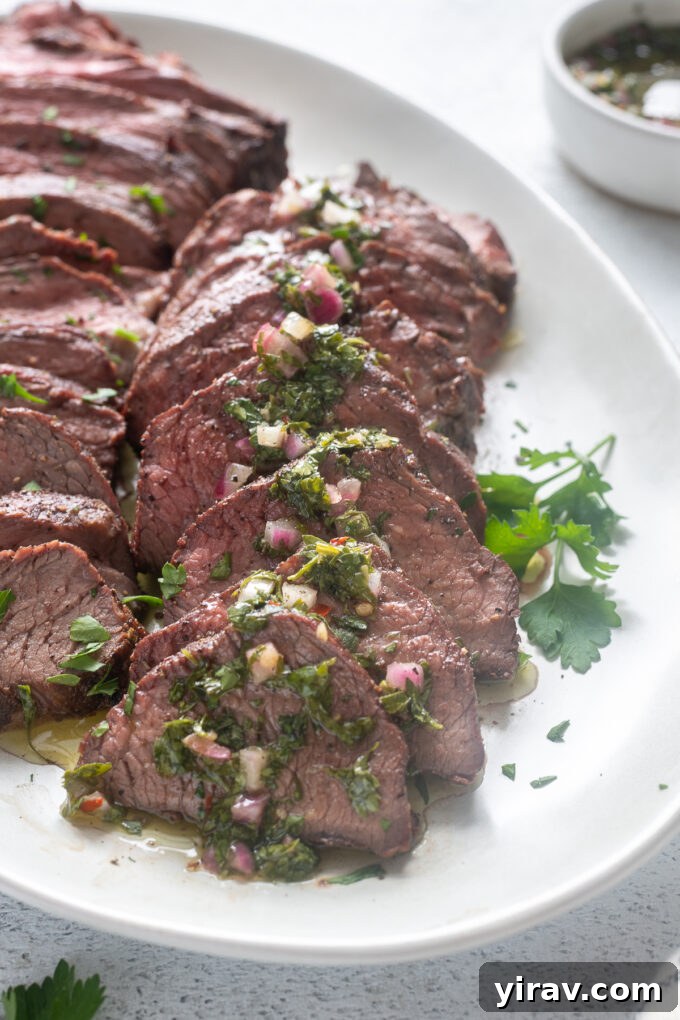 Grilled tri tip roast sliced on a platter, beautifully drizzled with chimichurri sauce and garnished with herbs.