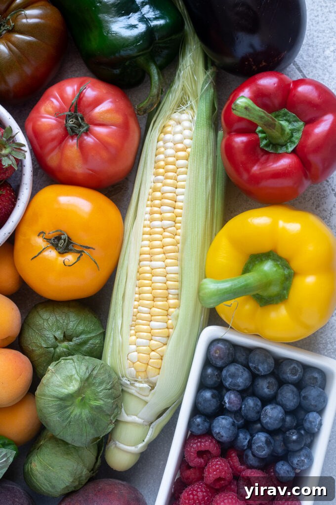 A beautiful flat lay of July seasonal fruits and vegetables, including golden corn, a variety of colorful berries, lush green nightshade vegetables, and more summer produce.