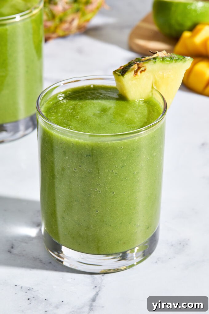 Tropical Green Power Smoothie 7 A glass of creamy mango kale smoothie with a vibrant green color, garnished with a pineapple wedge, on a rustic surface.