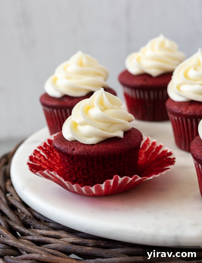 A perfectly baked red velvet cupcake with its paper liner partially removed, showcasing its moist, rich red interior and fluffy texture, topped with luscious cream cheese frosting.