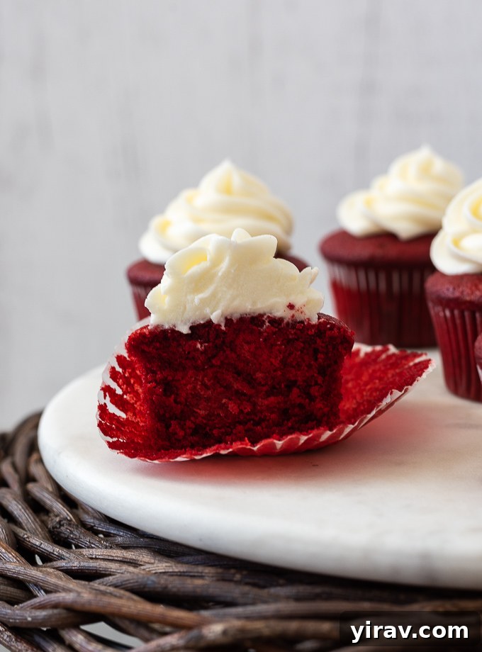 A red velvet cupcake perfectly cut in half, revealing its moist, open crumb texture and the beautiful vibrant red color, topped with a generous layer of cream cheese frosting.
