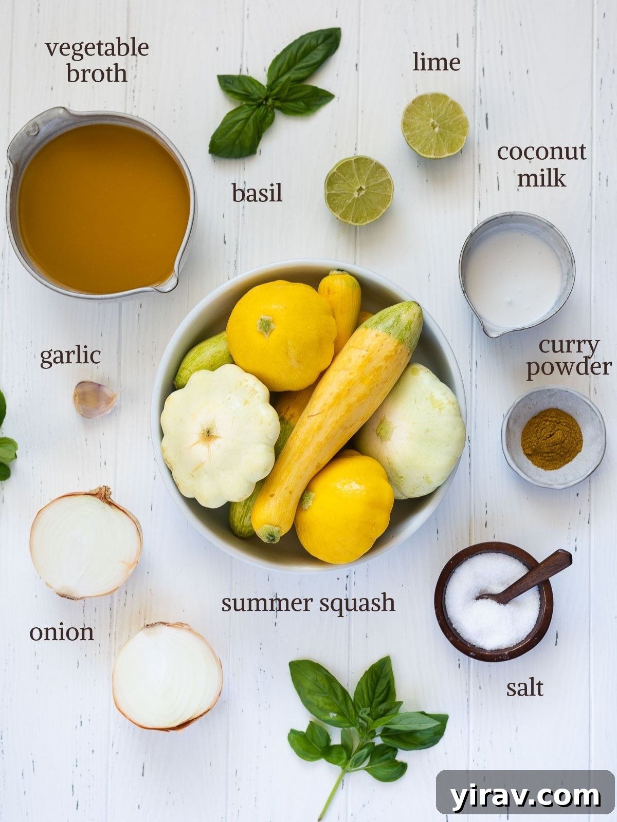 A flat lay of fresh ingredients for summer squash soup, including various types of yellow summer squash, a can of coconut milk, onions, garlic, fresh basil, and a bottle of vegetable broth.