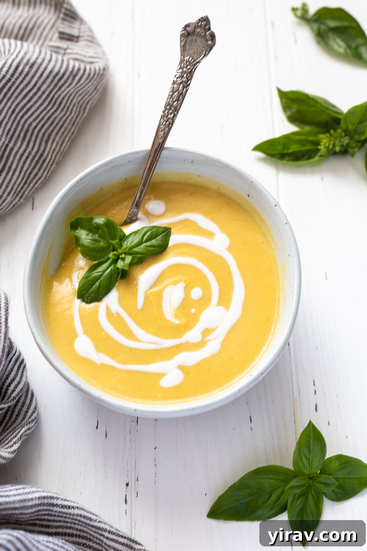 A beautifully presented bowl of creamy summer squash soup, garnished with fresh basil leaves. A silver spoon is placed in the bowl, ready to enjoy the rich texture.