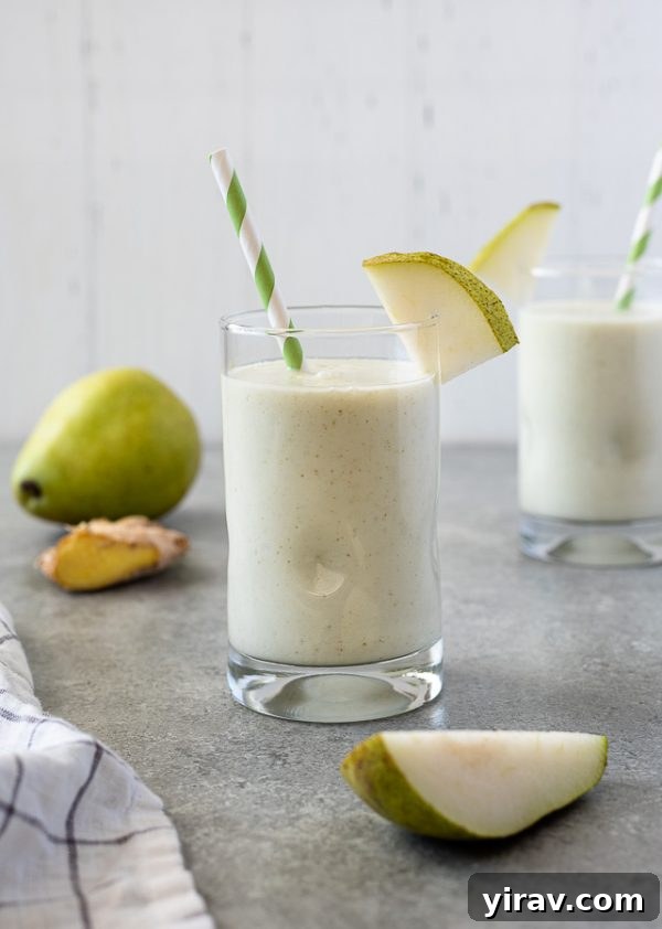 A tall glass of creamy pear ginger smoothie with a fresh pear slice on the rim and a green striped straw, hinting at a delightful, healthy drink.