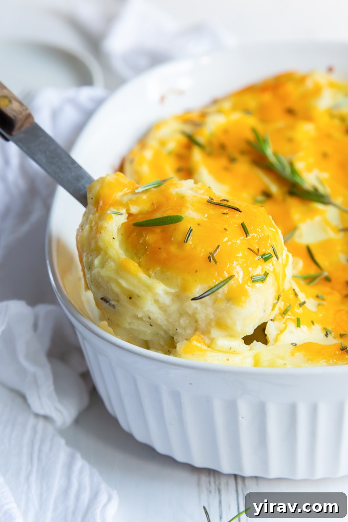 A serving spoon lifting a scoop of creamy, cheesy twice baked mashed potatoes out of a baking dish, garnished with fresh rosemary.