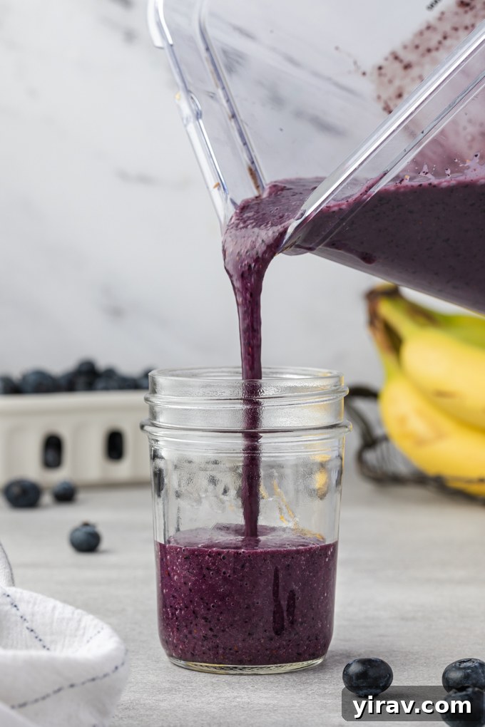 Thick, purple blueberry banana smoothie being poured from a blender into a clear mason jar, showcasing its rich texture and vibrant color.