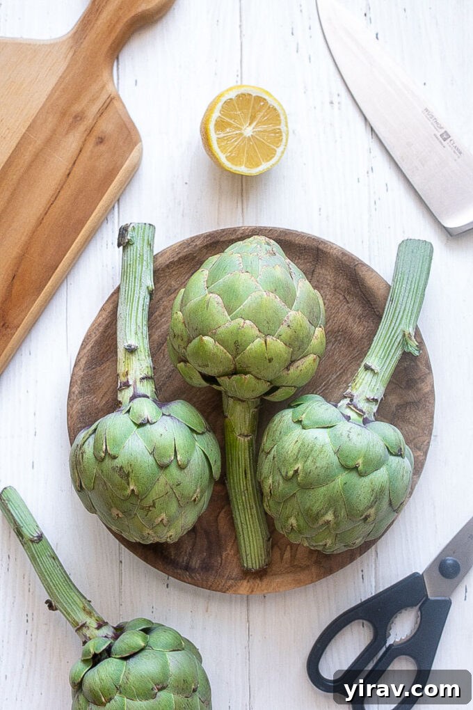Artichokes with knife lemon and tools for prepping on cutting board.